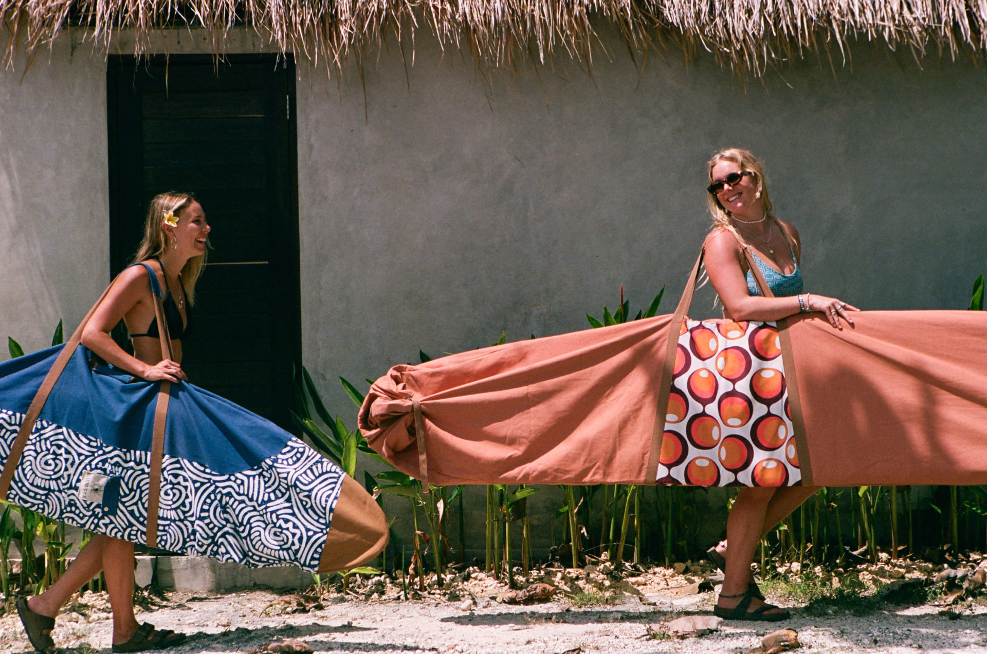 Two women carrying patterned Barefeet Surf board bags outside a tropical villa