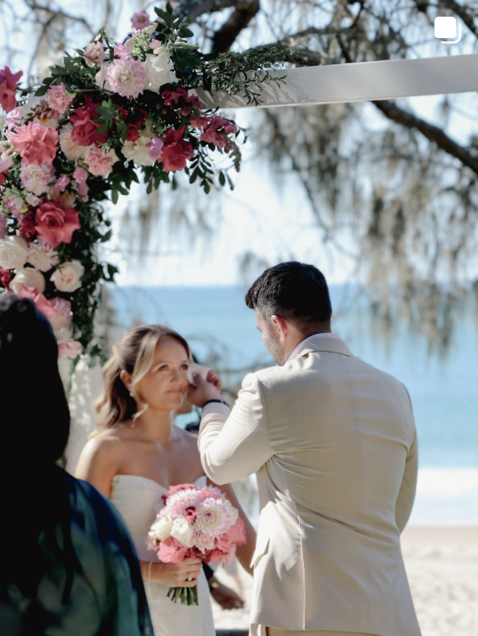 Beach wedding floral arch with pink roses as the couple exchanges rings